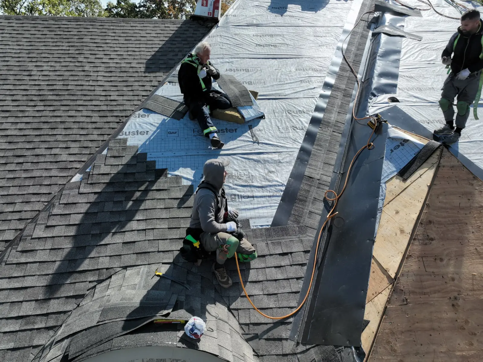 Roofers fastening grey shingles beside silver underlayment and exposed plywood on an Oakville roof