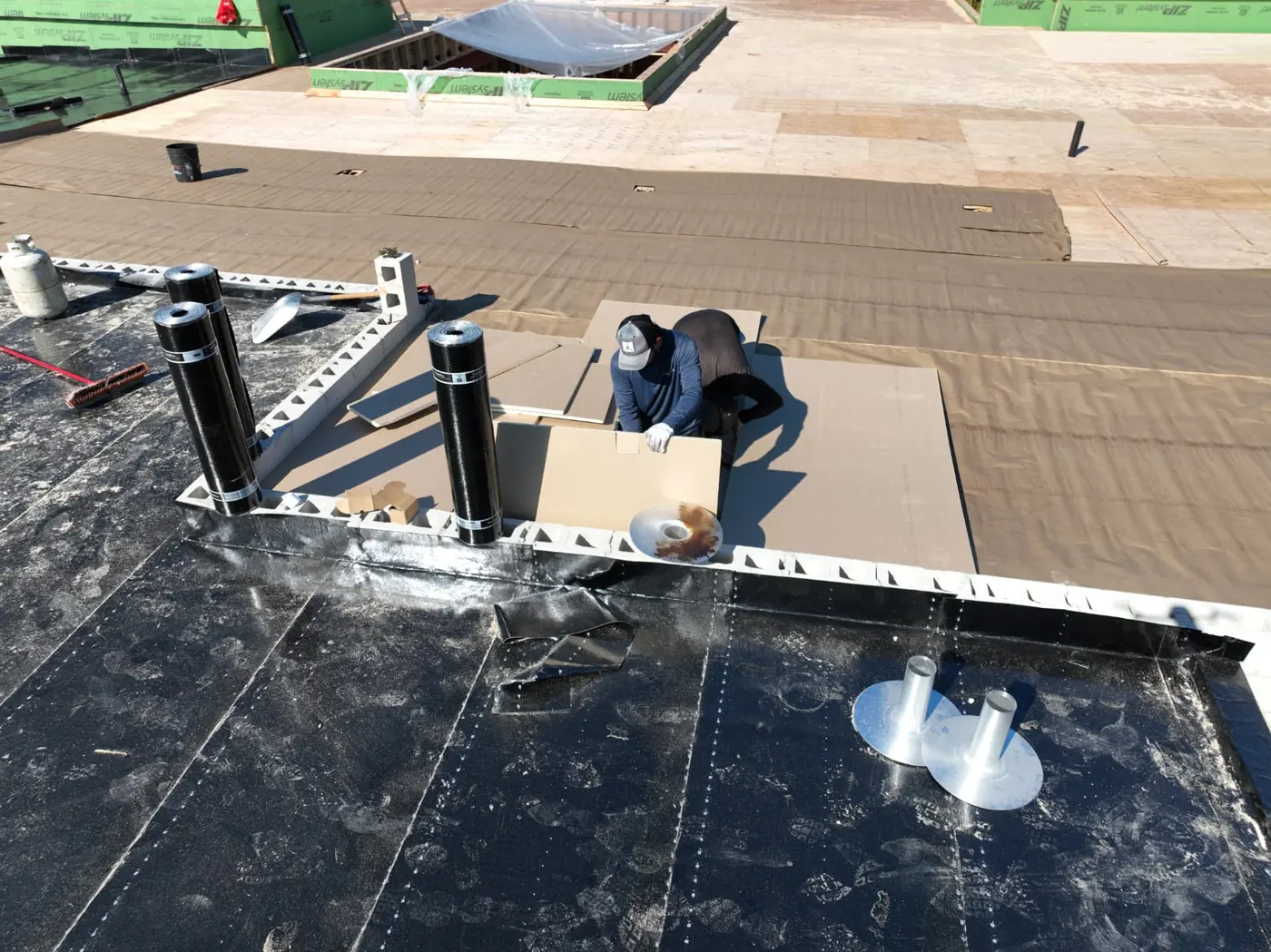 Roofer positioning rigid roof boards beside rolls of black membrane on a Brampton flat roof, with exposed plywood decking behind
