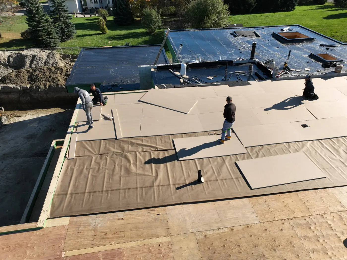 Wide aerial view of crew members installing rigid roof boards on a Brampton building, with black membrane sections, pipe openings and exposed plywood in the foreground