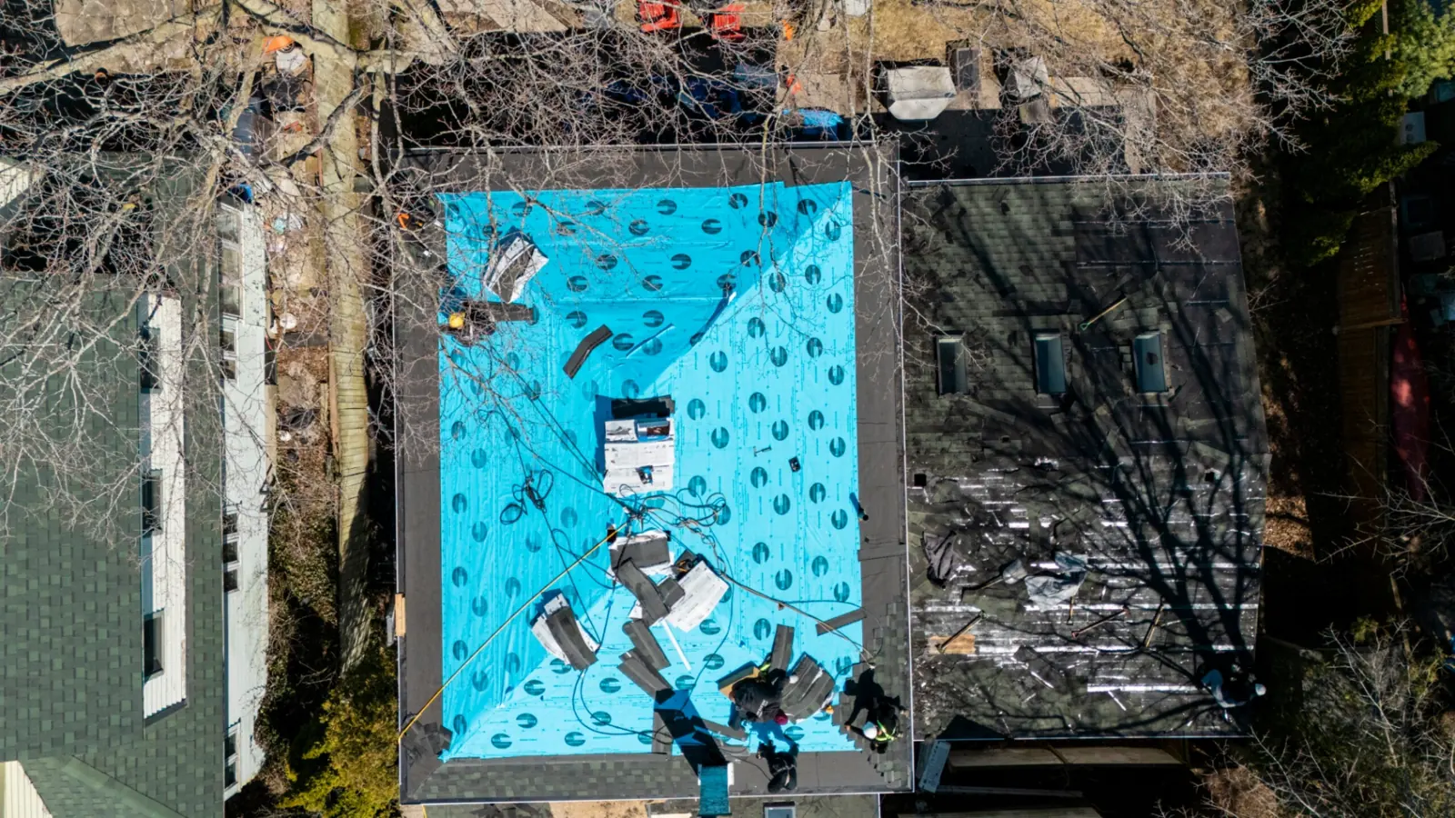 Overhead drone view of a roof covered in blue synthetic underlayment, with workers and a ladder