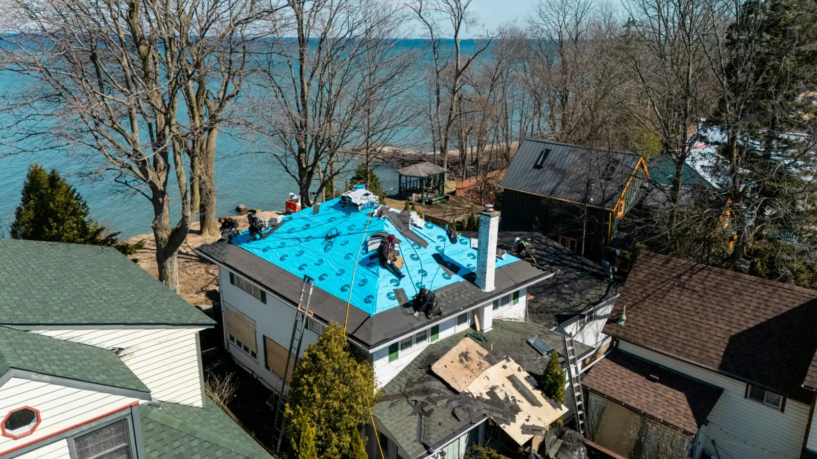 Aerial view of workers installing blue roof underlayment on a house beside trees and neighbouring homes