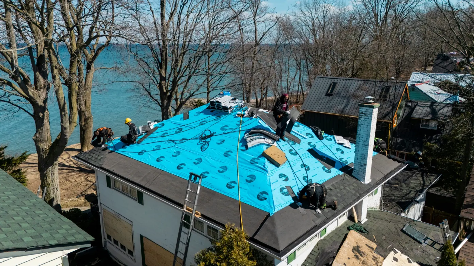 Drone view of a roof with blue synthetic underlayment, workers, a chimney and ladders along the eaves