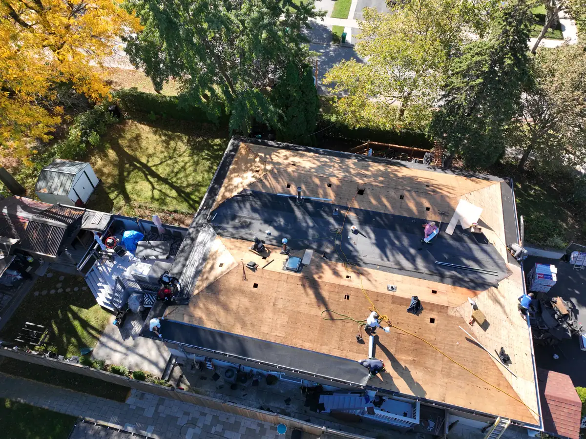Top-down drone view of a Mississauga hip roof stripped to the decking, with crew members installing ice and water shield along every edge