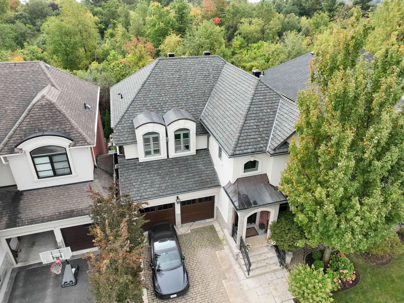 Aerial drone view of a finished Oakville roof with dark textured shingles, arched dormers and a metal porch canopy, with trees behind the property